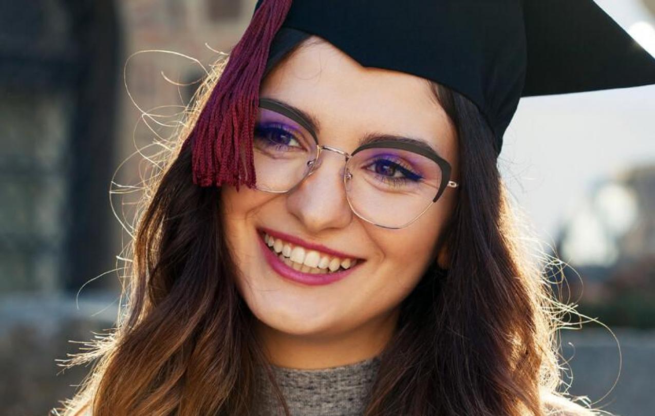 Photo of a girl with glasses in a cap and gown smiling. 